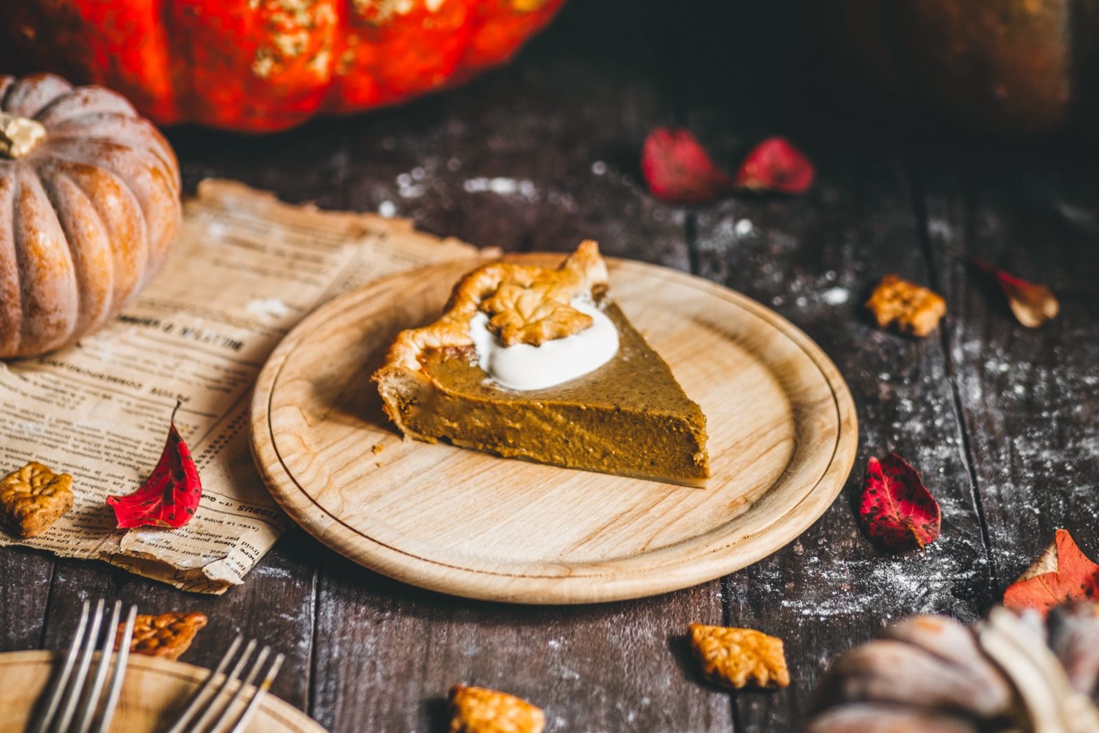 A slice of pumpkin pie topped with whipped cream, surrounded by leaves and pumpkins on a rustic tabletop.