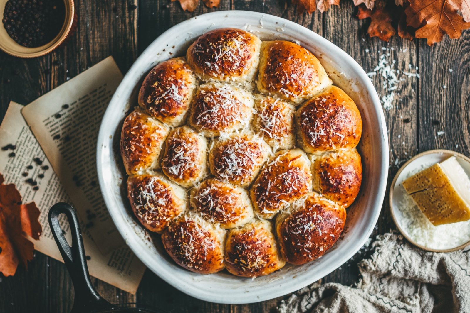 A dish of dinner rolls sprinkled with parmesan and black pepper, styled in a rustic and autumnal setting.