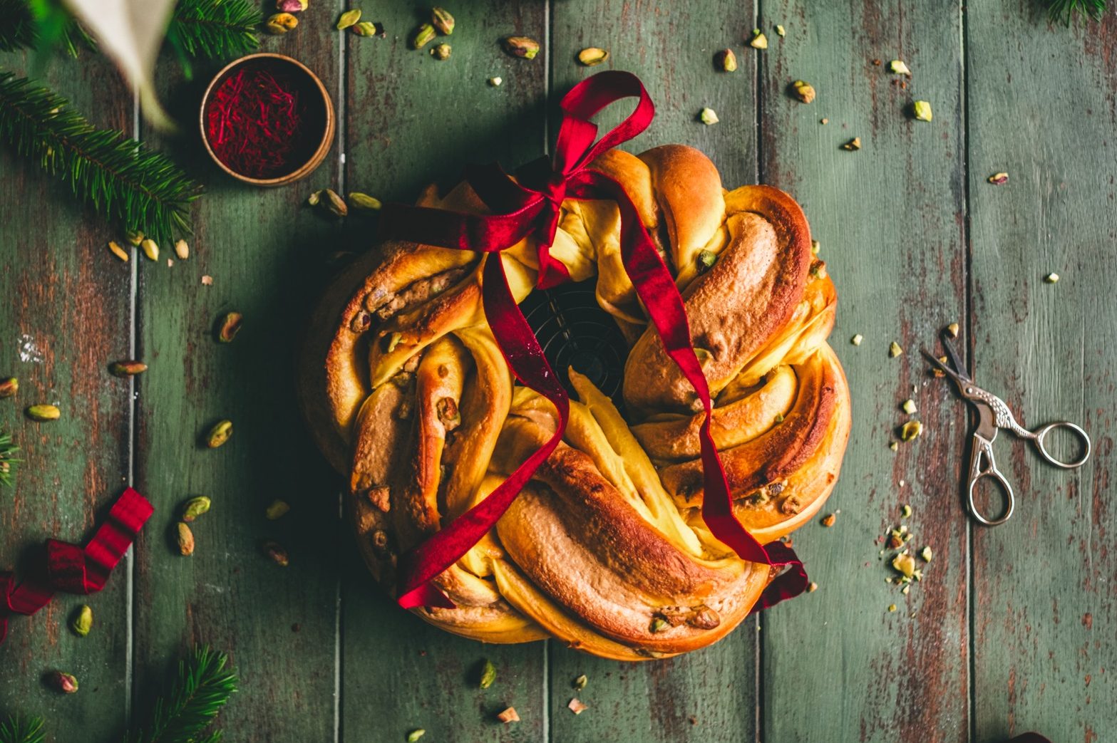A saffron and pistachio babka wreath styled on a rustic green backdrop. Pistachios are scattered around, and the scene is styled with saffron and holiday accents.