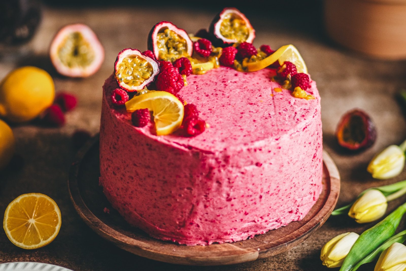 A lemon passionfruit cake with raspberry mascarpone buttercream styled in a bright but rustic setting. There are yellow tulips, sliced lemons, passionfruit, and raspberries that top the cake and are scattered around the scene.